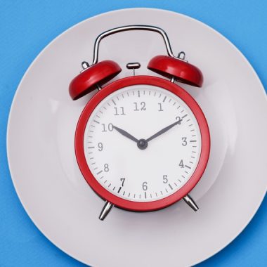 Red alarm clock lies on plate next to fork and knife